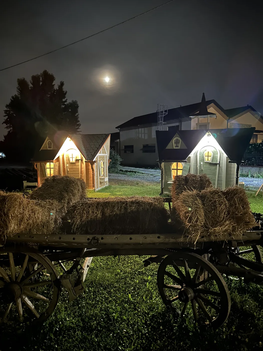 Casette fiabesche illuminate di notte sotto un antico carro agricolo con ruote in legno.