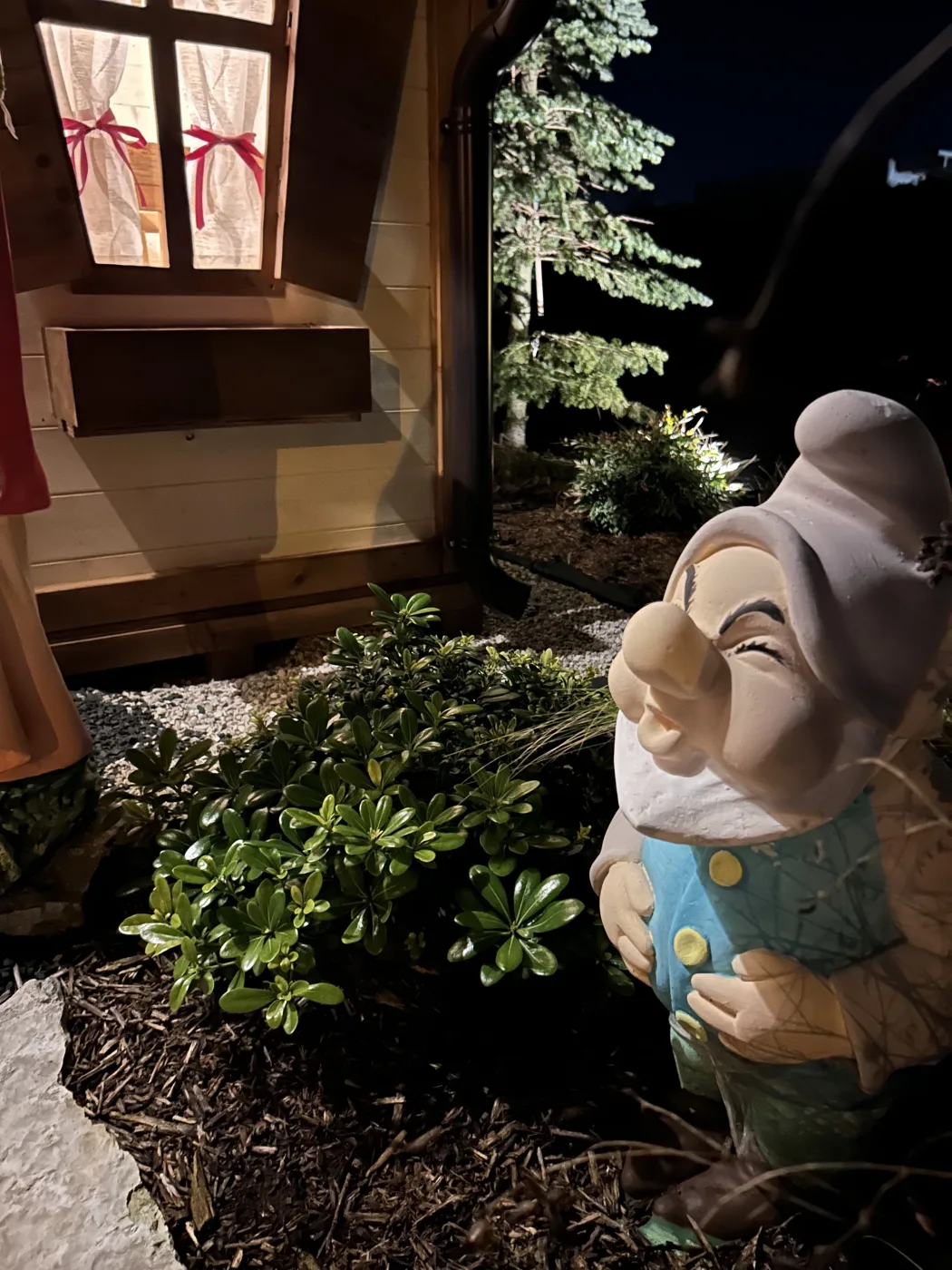 Statua del nano Pisolo nel giardino notturno illuminato davanti alla casetta.