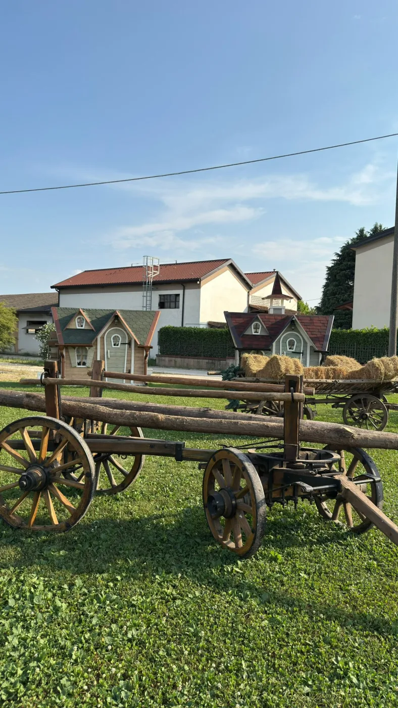 Casetta con doppio timpano e tetto verde a scandole vicino a un antico carro agricolo con una balla di fieno e una ghirlanda di pino.