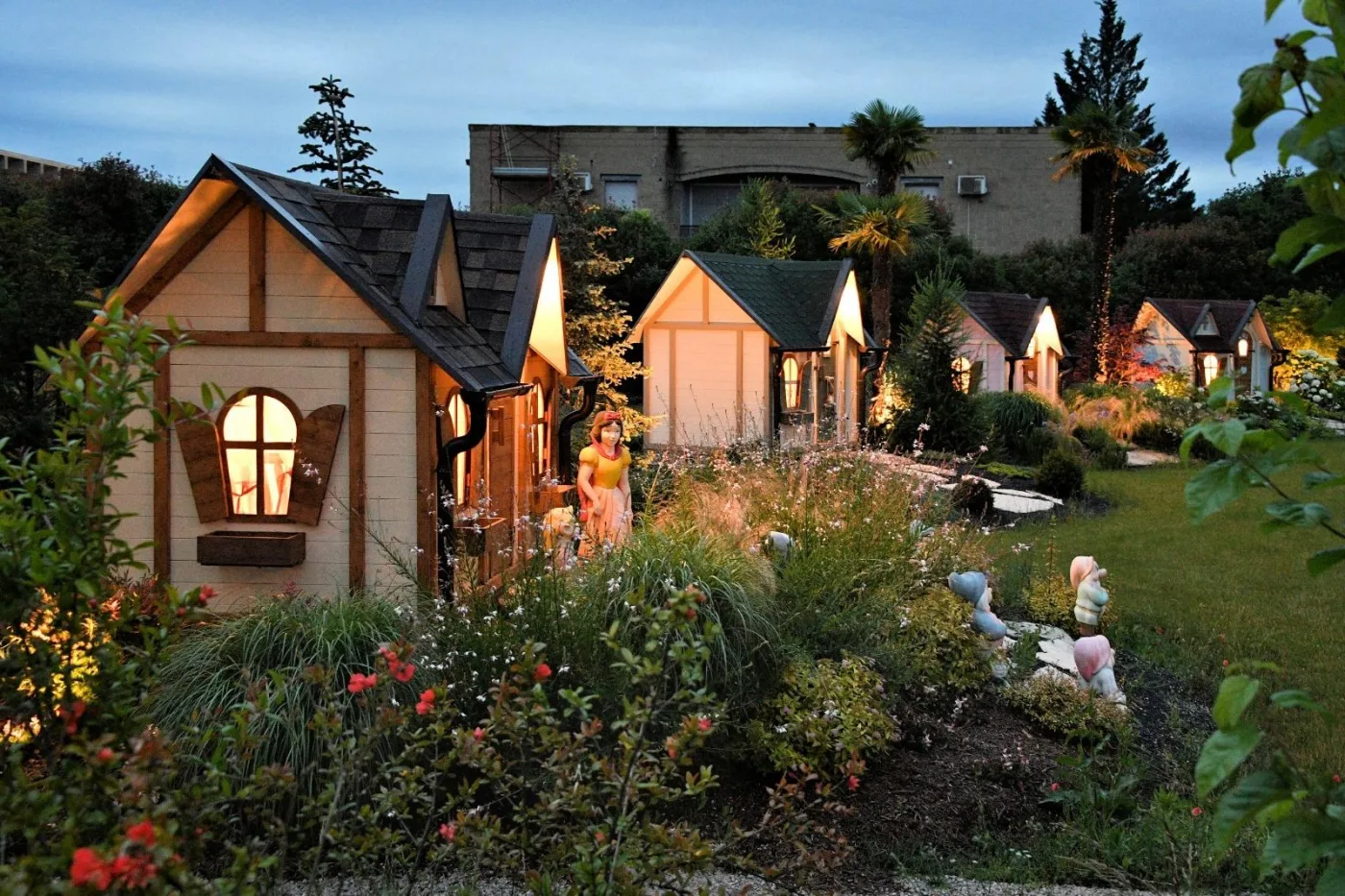 Fila di casette fiabesche illuminate al crepuscolo lungo il vialetto del giardino, con le statue di Biancaneve e dei sette nani in primo piano.